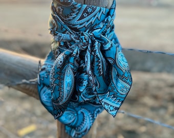 Peut inclure: Un bandana carré arborant un motif cachemire détaillé dans des tons bleu sarcelle, bleu clair, noir et blanc. Le tissu est noué autour d'un poteau en bois, révélant des motifs tourbillonnants complexes et de petites formes en étoile.