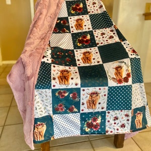 May include: A patchwork quilt with a teal background featuring a variety of floral and highland cow designs. The quilt is folded and draped over a chair with a pink faux fur throw.