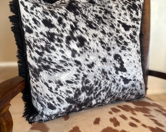 May include: A black and white cowhide pillow with a fringed border sits on a brown and white cowhide chair.