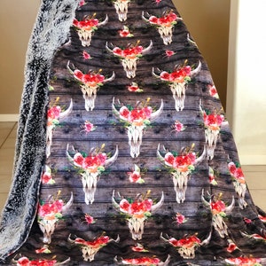 May include: A soft, plush throw blanket with a faux fur backing. The front features a repeating pattern of floral skull designs on a dark wood background.