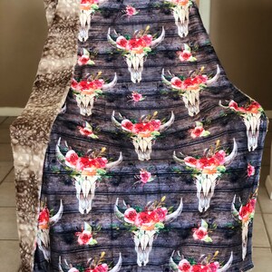 May include: A soft, plush throw blanket with a reversible design. One side features a floral skull pattern on a woodgrain background. The other side is a solid, light brown faux fur.
