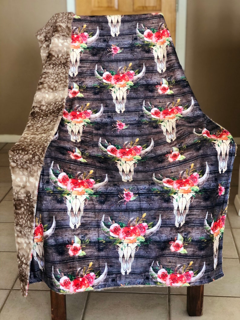 May include: A soft, plush throw blanket with a reversible design. One side features a floral skull pattern on a woodgrain background. The other side is a solid, light brown faux fur.