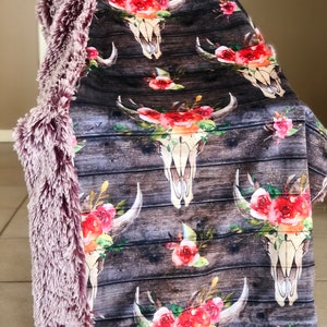 May include: A soft, plush throw blanket with a faux fur backing. The front side features a repeating pattern of watercolor floral designs and cow skulls on a woodgrain background.
