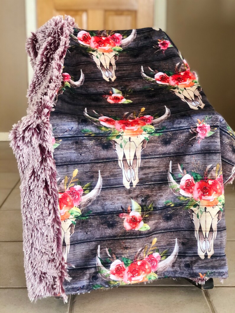 May include: A soft, plush throw blanket with a faux fur backing. The front side features a repeating pattern of watercolor floral designs and cow skulls on a woodgrain background.
