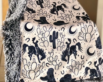 May include: A cream-colored blanket with a black and white pattern of cacti, horses, cowboy boots, and stars. The blanket has a gray faux fur border.