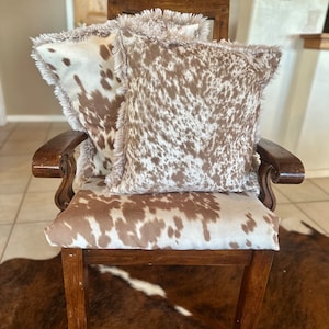 May include: A wooden chair with a brown and white cowhide patterned upholstery. The chair has two pillows with the same pattern and a fringe trim.