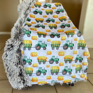 May include: A white fleece baby blanket with a gray faux fur border. The blanket features a repeating pattern of green tractors pulling hay bales.