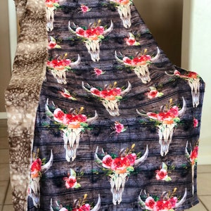 May include: A soft, plush throw blanket with a brown faux fur backing. The front features a repeating pattern of white skull designs with red and pink floral accents on a dark wood background.