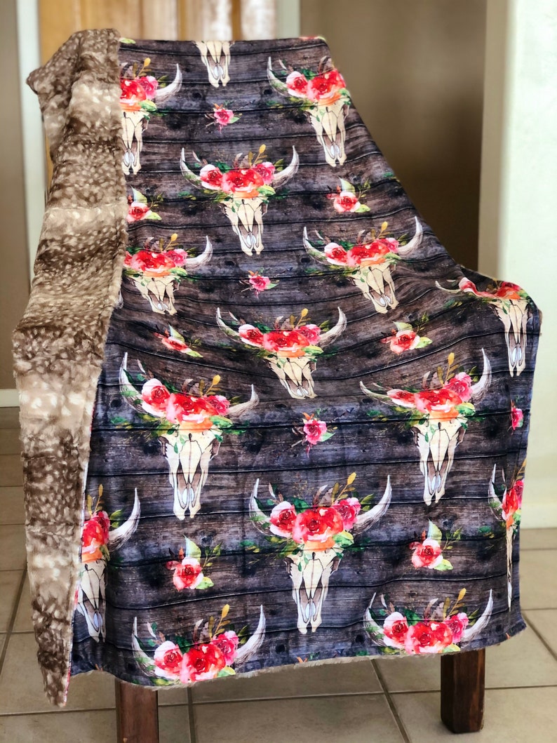 May include: A soft, plush throw blanket with a brown faux fur backing. The front features a repeating pattern of white skull designs with red and pink floral accents on a dark wood background.