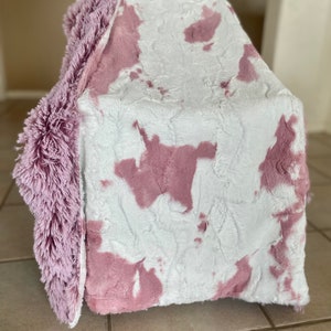 May include: A soft, plush throw blanket with a white and pink cow print design. The blanket has a fluffy, shaggy texture and a solid pink border.