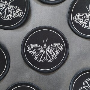 May include: Six black round patches with white embroidered butterfly designs. The butterflies are all facing the same direction with their wings spread out.