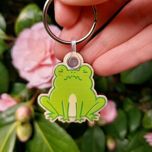 May include: A wooden frog keychain with a light brown background. The frog is green with a light brown belly and a small, white mouth. The keychain is attached to a silver key ring.