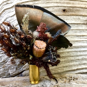 May include: A handcrafted boutonniere featuring a large brown feather, dried flowers, and a gold-colored bullet casing. The arrangement includes various textures and shades of brown, beige, and black, creating a rustic and natural aesthetic.