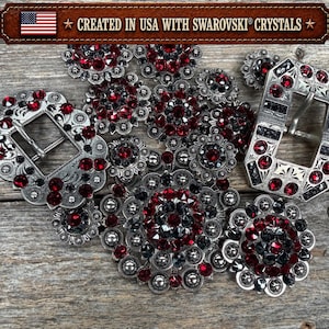 May include: A collection of silver-toned metal belt buckles and circular conchos embellished with deep red and black Swarovski crystals. The items feature intricate engraved patterns and are labelled with the text CREATED IN USA WITH SWAROVSKI CRYSTALS.