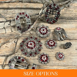 May include: A variety of silver-toned belt buckles and conchos with red rhinestone embellishments.  The buckles and conchos are arranged on a wooden surface.  SIZE OPTIONS