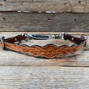 May include: A brown leather strap with a floral design and a silver snap hook. The strap is on a wooden surface.