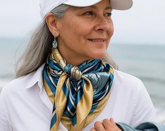 May include: A silk scarf with a floral and geometric design in shades of blue, gold, and white is knotted around the neck. The scarf is paired with a white button-down shirt and a white baseball cap. The woman is also wearing a pair of earrings.