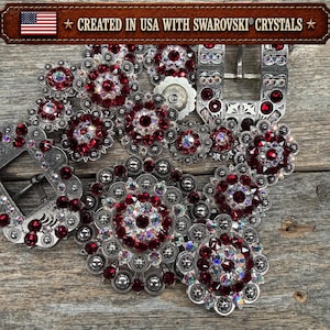 May include: A silver-toned belt buckle and matching concho set featuring deep red and iridescent Swarovski crystals. The metal components are circular and rectangular with intricate engraved detailing. Text at the top reads: CREATED IN USA WITH SWAROVSKI CRYSTALS.