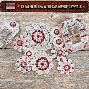 May include: A silver-toned belt buckle set featuring intricate floral-inspired metalwork adorned with deep red and iridescent Swarovski crystals. The set includes a large central buckle and a smaller matching buckle, both labeled Created in USA with Swarovski Crystals.