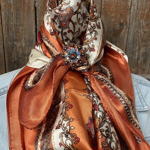 Op de afbeelding: Een bruine en witte paisley-gepatroneerde zijden sjaal met een grote, kleurrijke strass broche.