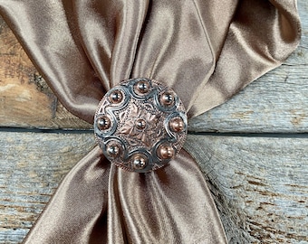 May include: A decorative bronze-colored scarf ring with a detailed floral and dot design. The ring is made of metal and is set against a draped, satin-like fabric in a matching bronze hue. The background is a rustic wooden surface.