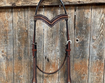 Può includere: Un morso per cavallo in pelle marrone con cuciture arancioni. Il morso è appeso a una recinzione di legno.