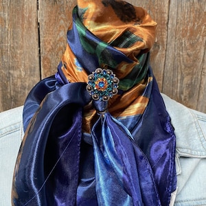 Op de afbeelding: Een blauwe en oranje zijden sjaal met een broche met juwelen in het midden. De sjaal is om de nek geknoopt.