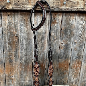 May include: A brown leather horse headstall with a floral design. The headstall has two buckles and is stitched with white thread.