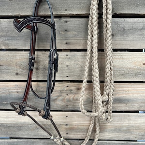 Puede incluir: Un cabestro de cuero marrón para caballos con costuras decorativas y una cuerda a juego. El cabestro presenta un color marrón oscuro con costuras más claras y herrajes plateados. La cuerda es de color beige claro y trenzada.
