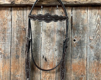 May include: A brown leather horse headstall with floral tooling and silver accents. The headstall is hanging on a wooden fence.