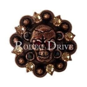May include: A copper-colored skull-shaped concho with the text "RODEO DRIVE" in white. The concho is surrounded by a decorative border with amber-colored and black gemstones.