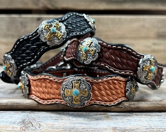 May include: Three leather dog collars with a woven pattern and silver cross accents. Each collar has a turquoise stone in the center of the cross. The collars are stacked on top of each other, with the black collar on top, the brown collar in the middle, and the tan collar on the bottom.