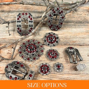 May include: Assortment of silver-toned belt buckles and conchos with red and black rhinestones. The buckles are in various shapes and sizes, including round, oval, and rectangular.