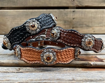 May include: Three leather dog collars with woven patterns and decorative metal accents featuring rhinestones. The collars are in black, brown, and tan.