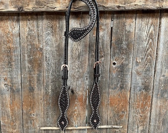 May include: A brown leather horse headstall with a decorative pattern and silver studs. The headstall is hanging on a wooden fence.