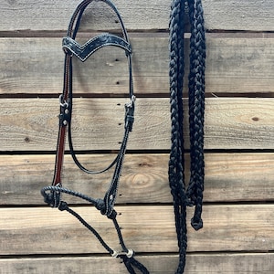 Könnte beinhalten: Ein schwarzer Lederpferdehalfter mit weißen Nähten und einem passenden schwarzen geflochtenen Seil. Der Halfter hat ein dekoratives geprägtes Design auf dem Stirnband und eine braune Lederunterlage.