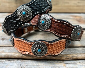 May include: Three leather dog collars with a woven pattern and silver flower-shaped buckles with turquoise stones. The collars are stacked on top of each other, with the black collar on the bottom, the brown collar in the middle, and the tan collar on top.