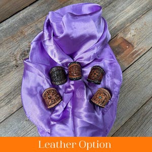 May include: Five leather bandana slides with different designs and colors, including brown, black, and tan. The slides are arranged on a purple satin fabric.