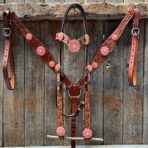 May include: A brown leather horse tack set with pink daisy accents. The set includes a headstall, breast collar, and reins. The leather is tooled with floral patterns and features silver-tone hardware. The background is a rustic wooden wall.
