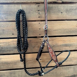 May include: A brown leather horse halter with silver accents and a black braided rope. The halter has a floral design and is hanging on a wooden wall.