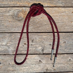 May include: A red rhinestone bolo tie with silver tips. The tie is knotted and laid out on a wooden surface.