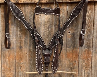 Puede incluir: Un juego de guarnicionería de caballo de cuero marrón con costuras blancas. El juego incluye un cabezal, un pecho y riendas.