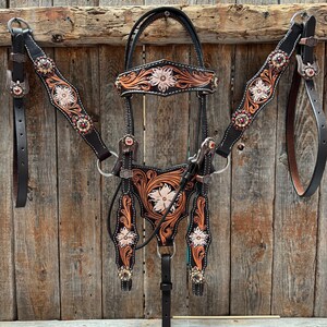 May include: A black and brown leather horse tack set with floral tooling and jewel accents. The set includes a bridle, breast collar, and reins, all with intricate designs and decorative elements. The background is a rustic wooden wall.