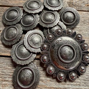 May include: A collection of silver-toned metal conchos featuring intricate floral engravings and a rope-style border. The set includes several circular conchos and one larger, sunburst-shaped concho with small silver spheres along the outer edge.