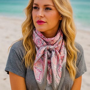 May include: A pink and gray paisley patterned scarf tied around the neck. The scarf has a square shape with a gray border and is made of a soft, flowing material. The scarf is worn over a gray t-shirt.