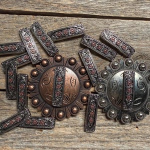 May include: A collection of metal hardware including two large circular conchos and several rectangular bars. The pieces feature silver and copper-toned finishes with embossed floral patterns and small circular studs along the edges.