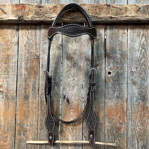 May include: A brown leather horse headstall with white stitching and silver hardware. The headstall is hanging on a wooden fence.