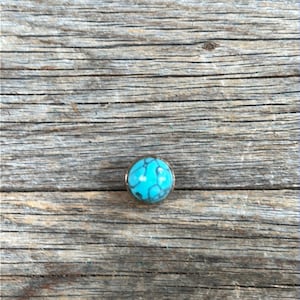 May include: A turquoise blue cabochon stone set in a silver tone metal setting. The stone has a marbled pattern.