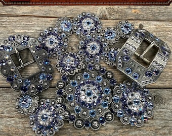 May include: Two silver-toned metal belt buckles and several circular concho ornaments, all embellished with blue and clear Swarovski crystals. The items feature intricate engraved patterns and are arranged together in a cluster.
