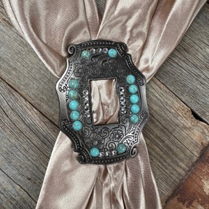 May include: A silver-toned metal bolo tie slide with an ornate design and turquoise-colored stones. The slide is set against a light brown fabric background.
